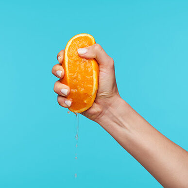 Indoor photo of a hand holding orange and clenching a fist while squeezing juice, preparing breakfast while posing over blue background