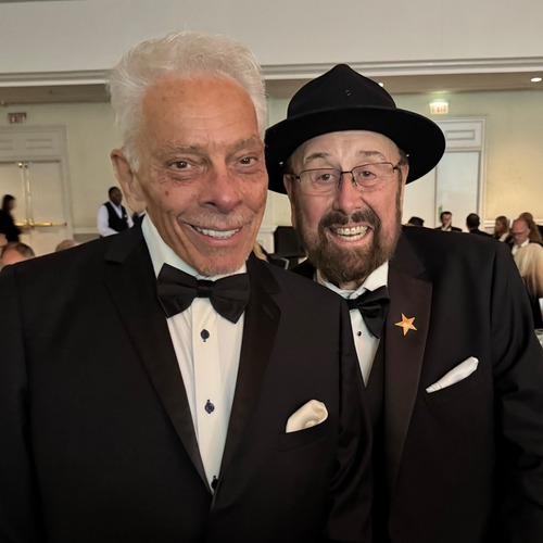 Two men in tuxedos smile at the camera at a banquet